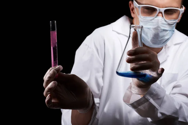 Assistant de laboratoire dans un masque médical et dans un manteau blanc avec un liquide dans une fiole et un produit chimique sur un fond noir — Photo