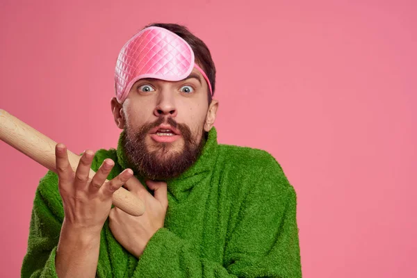 Retrato de un hombre con una máscara de sueño rosa y un rodillo de madera emociones modelo de irritabilidad bata verde — Foto de Stock