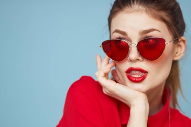 Koyu gözlükleri olan güzel bir kadın parlak makyajlı kırmızı dudaklar mavi arkaplanı büyülüyor.