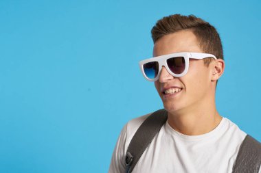 Kotlu, dizleri yırtık rahat bir adam mavi arka planda bir sandalyede oturuyor ve yüzünde uzun gözlüklerle oturuyor.