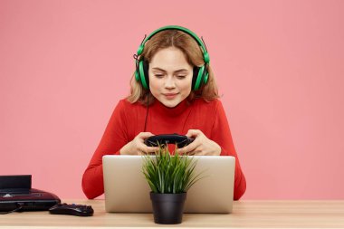 Kulaklık takan, dizüstü bilgisayarlı bir kadın masa başı eğlence sektöründe pembe arka planda oturuyor.