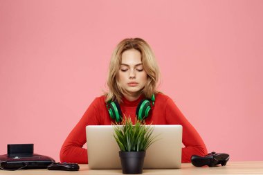 Kulaklık takan, dizüstü bilgisayarlı bir kadın masa başı eğlence sektöründe pembe arka planda oturuyor.