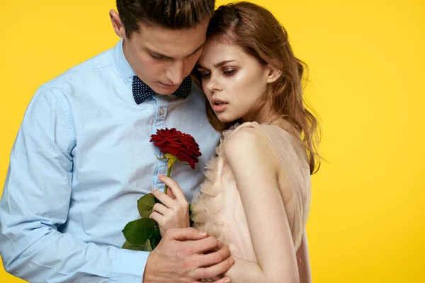 A loving young couple with a red rose in their hands are hugging on a yellow background — Stock Photo, Image