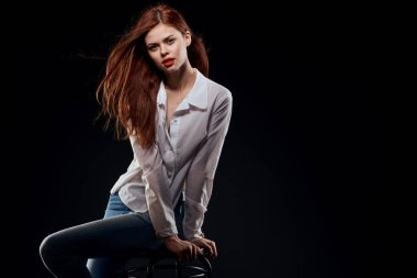 Sandalyede oturan güzel bir kadın beyaz gömlek, kot pantolon, uzun saç, kırmızı dudaklar koyu arka plan.