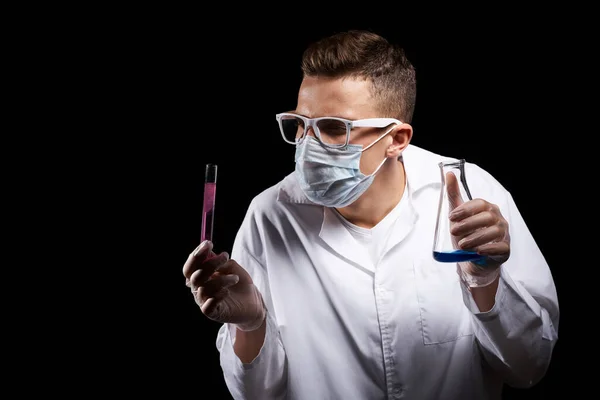 Лабораторний асистент у медичній масці та в білій шубці з рідиною в колбі та хіміком на чорному тлі — стокове фото