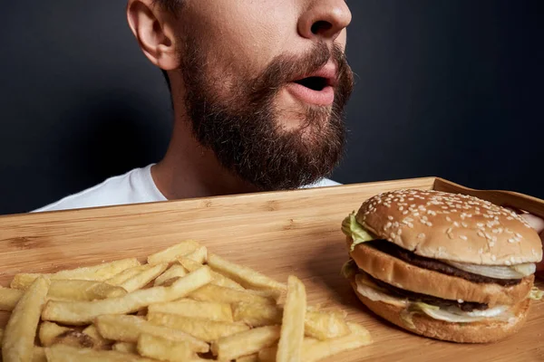 Emotivní muž s dřevěnou paletou rychlé občerstvení hamburger hranolky jíst jídlo životní styl tmavé pozadí — Stock fotografie