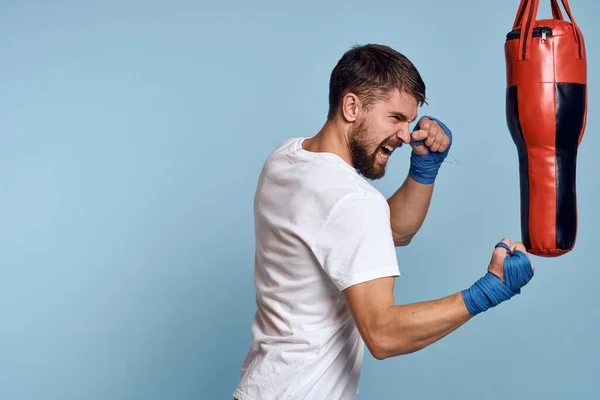 Un homme pratiquant un coup de poing sur un sac de boxe dans un t-shirt blanc sur un fond bleu — Photo