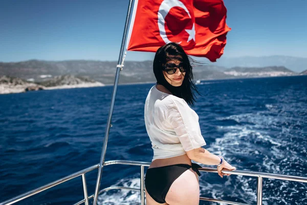 Slim woman in black bikini posing on yacht with Turkisf flag on sea background