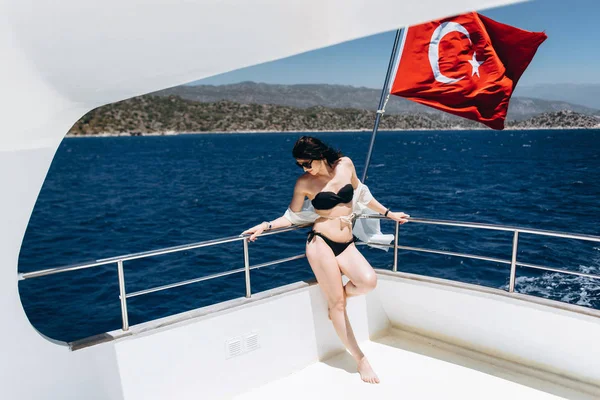 Slim woman in black bikini posing on yacht with Turkisf flag on sea background