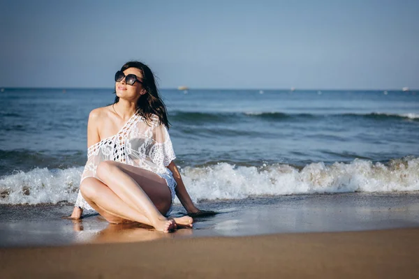 Attractive woman in sunglasses and bikini sitting on wet seashore