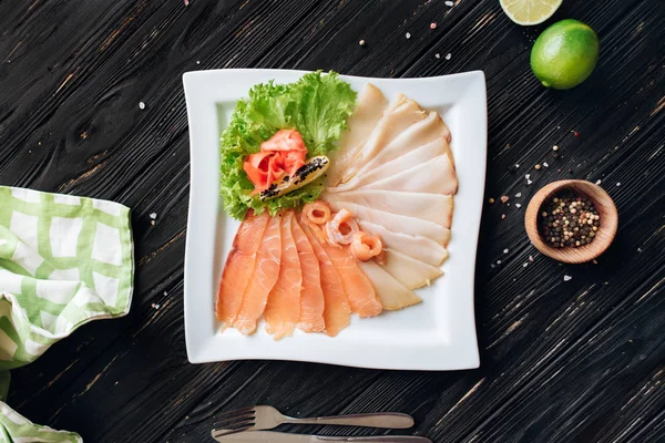 Peppper, limon, çatal bıçak takımı ve damalı yeşil peçete ile plaka üzerinde dilimlenmiş çeşitli kürlenmiş balık.