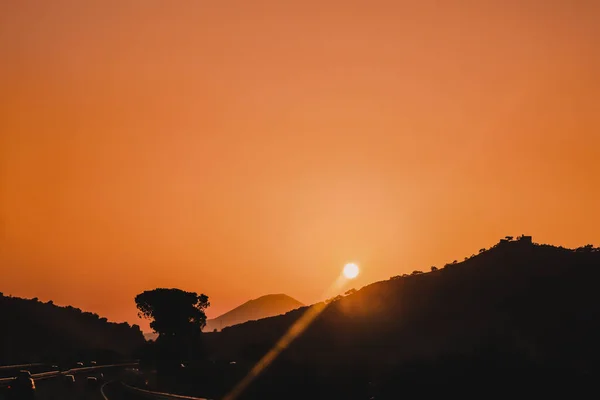Beautiful orange sky sunset on highway with cars driving around the mountains with sun above and cedar trees silhouettes. Summer dusk landscape of monti Lattari, Campania, Italy. Copy space
