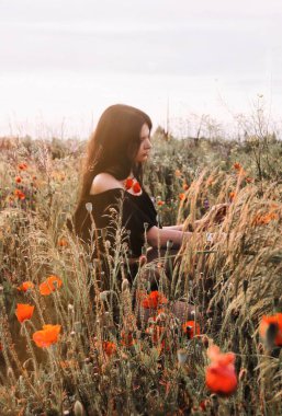 Doğayla konuşmak. Koyu kahverengi saçlı, omuzları açık, genç, güzel esmer bir kadın. Siyah bir elbise. Yaban gelincikleri çiçekleri ve sivri uçlu bir yaz tarlasında oturuyor.