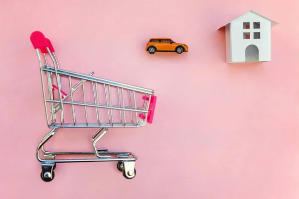 Small supermarket grocery push cart for shopping toy miniature white house and car on pink pastel color paper flat lay background. Mortgage property insurance buy dream home shopping sale rent concept