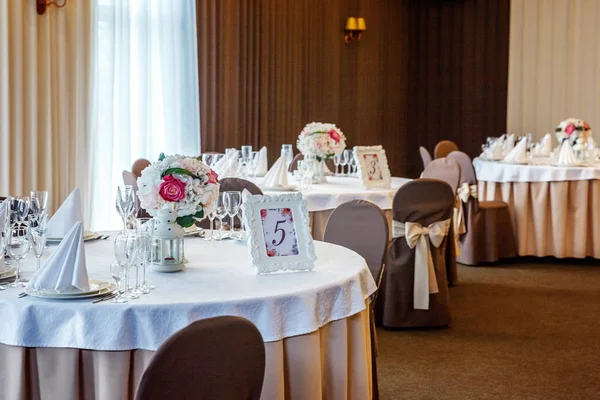 Glasses, flowers, fork, knife, napkin folded in a pyramid, served for dinner in restaurant with cozy interior. Wedding decorations and items for food, arranged by the catering service on a large table covered with white tablecloth