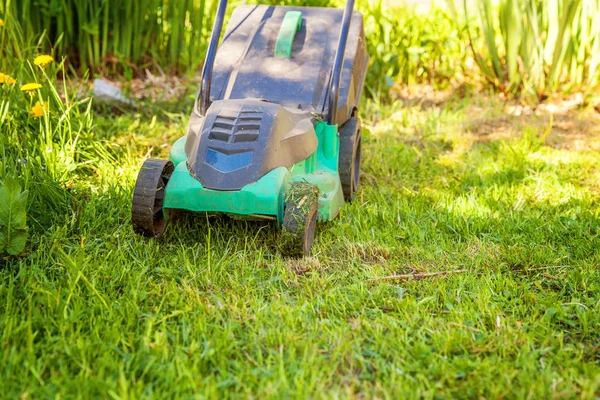 Lawn mower cutting green grass in backyard in sunny day. Gardening country lifestyle background. Beautiful view on fresh green grass lawn in sunlight, garden landscape in spring or summer season