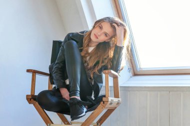 Deri ceketli beyaz duvar arka plan pencere karşı parlak ışık odada poz modern sandalye üzerinde oturan genç Genç kız. Hipster rock tarzı kıyafet giymiş seksi moda şehvetli kadın