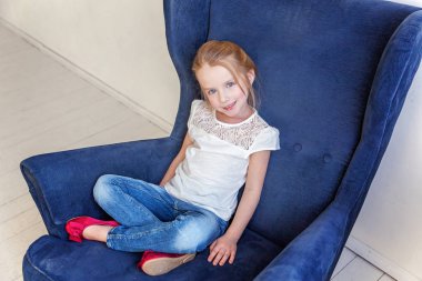 Tatlı küçük kız kot ve beyaz tişört evde modern rahat mavi sandalye beyaz oturma odasında rahatlatıcı üzerinde oturan