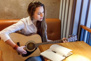 Young smiling hipster woman sitting playing guitar and write a song at cafe. Girl learning to play music clipart