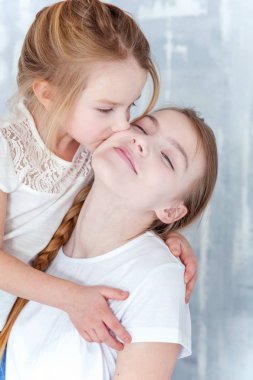 Gri desenli duvar arka plan karşı duran ve kucaklayan iki mutlu çocuk. Sevimli sevimli küçük kız onun sevgi ve bakım gösterilen sıkı Şirin genç kız, sarılma. Kız evde eğlenmek