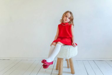 Mutlu küçük kız beyaz duvar arka planda parlak oda karşı sandalyeye oturan kırmızı elbiseli