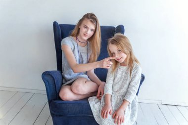 Modern rahat mavi sandalye evde beyaz oturma odasında rahatlatıcı oturan iki mutlu çocuk. Onun sevgi ve bakım gösterilen sevimli genç kız ile oynayan sevimli sevimli küçük kız. Kız evde eğlenmek