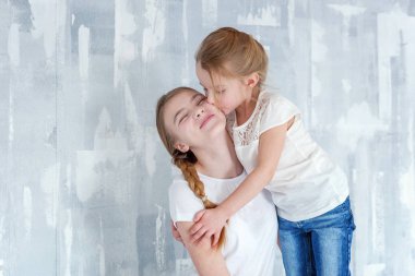 Gri desenli duvar arka plan karşı duran ve kucaklayan iki mutlu çocuk. Sevimli sevimli küçük kız onun sevgi ve bakım gösterilen sıkı Şirin genç kız, sarılma. Kız evde eğlenmek