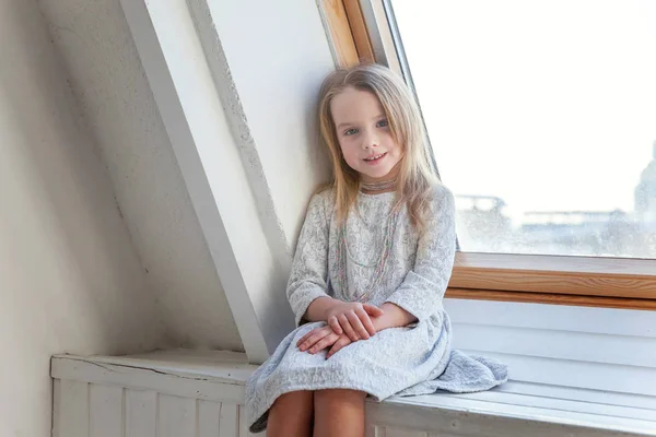 明るい光の居間で自宅の窓枠に座っていると考えて白いドレスでかわいい甘い笑顔女の子 — ストック写真