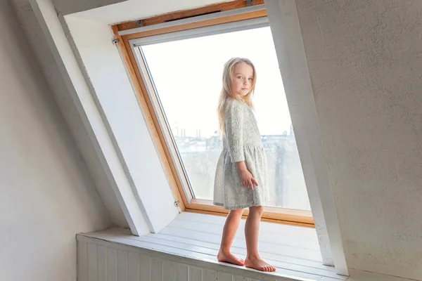 Little cute sweet smiling girl in white dress standing on the window sill in bright light living room at home and thinking