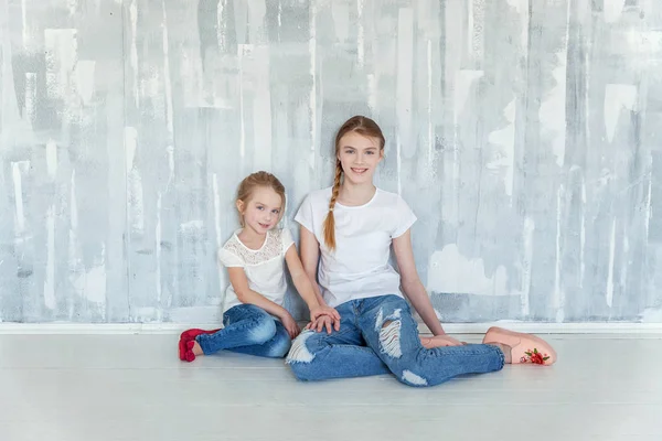 Two happy kids sitting against grey textured wall background and embracing. Adorable pretty little girl hugging tight cute teenage girl, showing her love and care. Sisters having fun at home