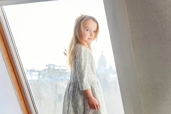 Little cute sweet smiling girl in white dress standing on the window sill in bright light living room at home and thinking