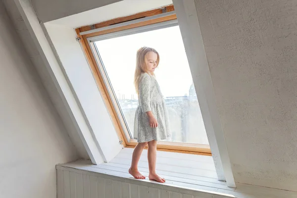 Stock image Little cute sweet smiling girl in white dress standing on the window sill in bright light living room at home and thinking