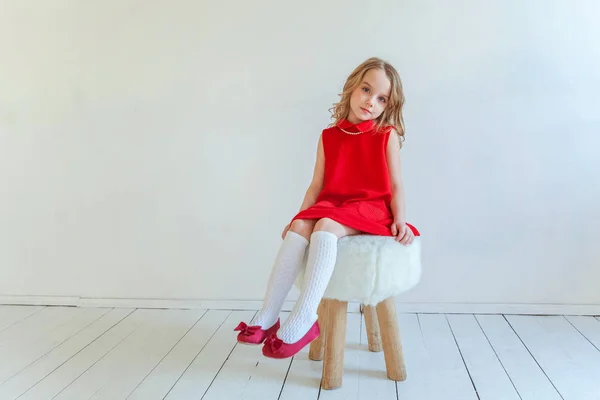 明るい部屋で白い壁背景に椅子に座っている赤いドレスの甘い幸せな少女 — ストック写真