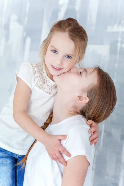 两个快乐的孩子站在灰色纹理的墙背景和拥抱 可爱的漂亮的小女孩拥抱紧可爱的少女 显示她的爱和关怀 姐妹们在家里玩得很开心 — 图库照片