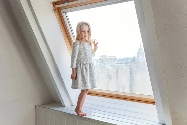 明るい光自宅居間の窓の土台の上に立って 考えて白いドレスでかわいい甘い笑顔女の子 — ストック写真
