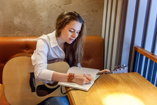 年轻的微笑的嬉皮士妇女坐在那里弹吉他 并在咖啡馆里写一首歌 女孩学习演奏音乐 — 图库照片