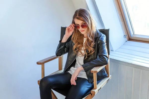 Young teenage girl in leather jacket sitting on modern chair posing in bright light room against white wall background window. Pretty sexy fashion sensual woman dressed in hipster rock style outfit
