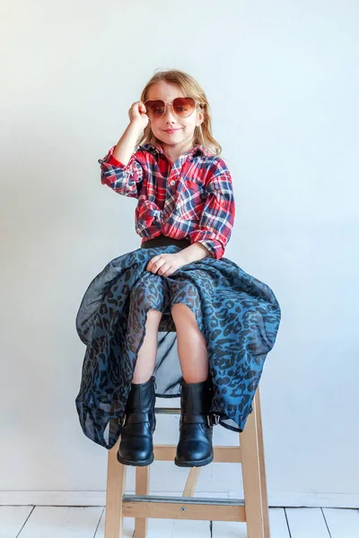 Sweet happy adorable fashion young little girl dressed like hipster hippie gipsy cowboy in sunglasses sitting on modern chair posing in white wall background in light bright room. Lifestyle people trendy concept