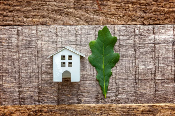 Miniature white toy model house with green oak leaf on wooden backgdrop. Eco Village, abstract environmental background. Real estate mortgage property insurance dream home ecology concept