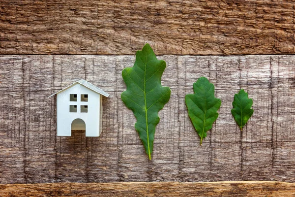 Miniature white toy model house with green oak leaves on wooden backgdrop. Eco Village, abstract environmental background. Real estate mortgage property insurance dream home ecology concept