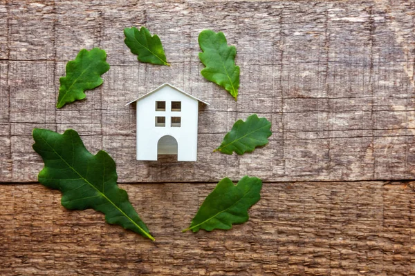 Miniature white toy model house with green oak leaves on wooden backgdrop. Eco Village, abstract environmental background. Real estate mortgage property insurance dream home ecology concept