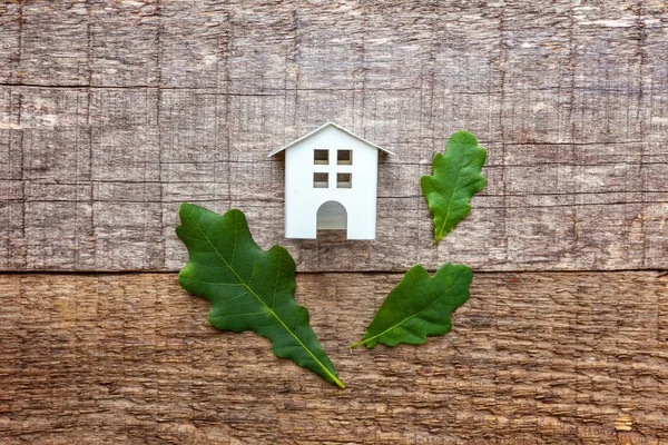 Miniature white toy model house with green oak leaves on wooden backgdrop. Eco Village, abstract environmental background. Real estate mortgage property insurance dream home ecology concept