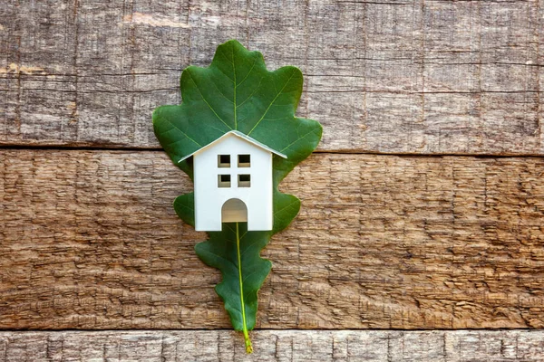 Miniature white toy model house with green oak leaf on wooden backgdrop. Eco Village, abstract environmental background. Real estate mortgage property insurance dream home ecology concept