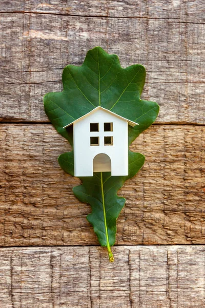 Miniature white toy model house with green oak leaf on wooden backgdrop. Eco Village, abstract environmental background. Real estate mortgage property insurance dream home ecology concept