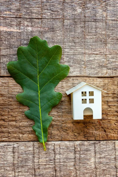Miniature white toy model house with green oak leaf on wooden backgdrop. Eco Village, abstract environmental background. Real estate mortgage property insurance dream home ecology concept