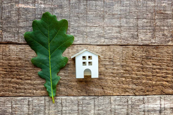 Miniature white toy model house with green oak leaf on wooden backgdrop. Eco Village, abstract environmental background. Real estate mortgage property insurance dream home ecology concept