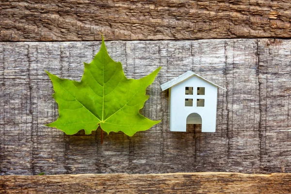 Miniature white toy model house with green maple leaf on wooden backgdrop. Eco Village, abstract environmental background. Real estate mortgage property insurance dream home ecology concept