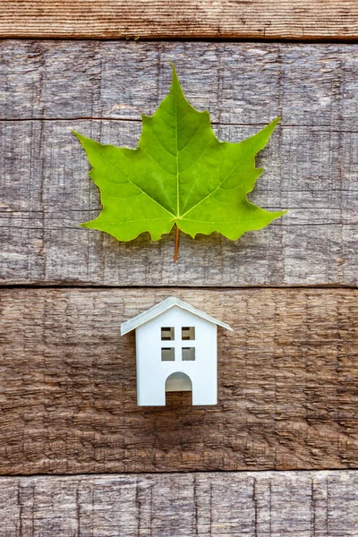 Miniature white toy model house with green maple leaf on wooden backgdrop. Eco Village, abstract environmental background. Real estate mortgage property insurance dream home ecology concept