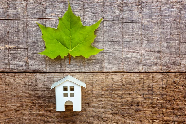 Miniature white toy model house with green maple leaf on wooden backgdrop. Eco Village, abstract environmental background. Real estate mortgage property insurance dream home ecology concept