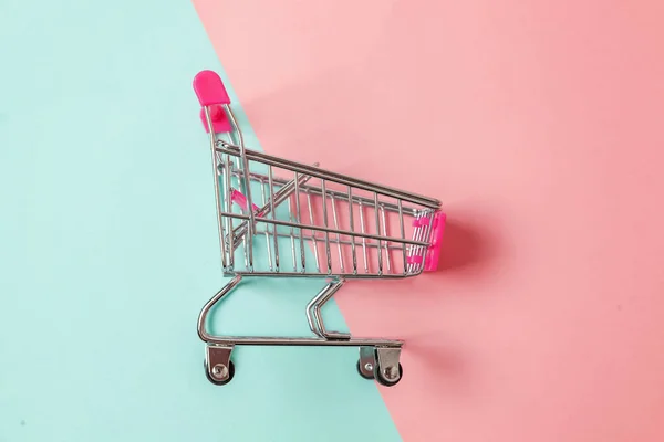 Small supermarket grocery push cart for shopping toy with wheels isolated on blue and pink pastel colorful paper trendy geometric background Copy space. Sale buy mall market shop consumer concept
